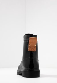 Bottines noires brillantes avec une semelle en caoutchouc texturé, une étiquette en cuir avec logo, et un dessus lisse avec des détails de couture subtils.