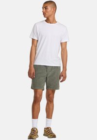Witte katoenen t-shirt, olijfgroene corduroy shorts, witte crew sokken en bruine sportieve schoenen met kleurrijke veters. Eenvoudige, casual outfit.