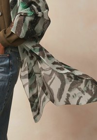 Sheer, pleated scarf with a green and black abstract pattern. Lightweight fabric drapes elegantly, creating a flowing texture.