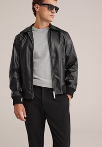 Svart läder bomberjacka med dragkedja framtill, ribbade manschetter och krage, lager på lager över en grå tröja och svarta byxor.