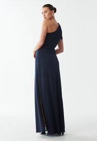 Abito maxi blu navy con spalla unica, tessuto drappeggiato, spacco laterale e vita aderente. Texture liscia con una silhouette che valorizza.