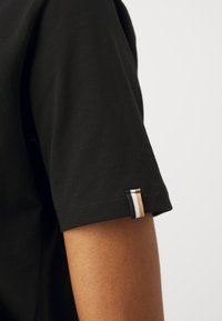 Black cotton t-shirt with short sleeves; features a small multi-colored label with black, white, and gold stripes at the sleeve hem.