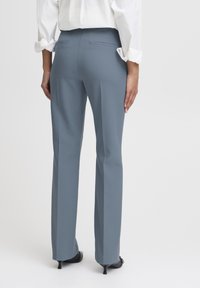 Op maat gemaakte, hoge broeken van een middelgrijze stof, met twee achterzakken en een rechte pijp, gecombineerd met zwarte hakken.