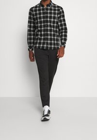 Camicia a quadri bianca e nera con colletto, abbinata a pantaloni scuri e scarpe da ginnastica bianche e nere, su uno sfondo neutro.