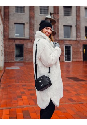 Weiße Kunstpelzjacke mit Schalkragen, schwarze Umhängetasche mit Logo, graue Handschuhe und schwarze Mütze, vor einem Ziegelmauer-Hintergrund.