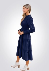 Vestido azul marino de volantes con mangas largas, cuerpo fruncido y puños con volantes. Llevado con botas blancas hasta el tobillo con tacón cuadrado.