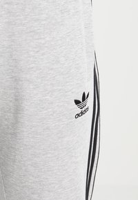 Grå sweatpants i mjukt material, med svarta sidostreck och en svart Adidas-logotyp på vänster lår.