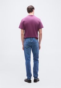 Άνδρας που στέκεται γυρισμένος με την πλάτη φορώντας μωβ κοντομάνικο πουκάμισο, μπλε τζιν παντελόνι και μαύρα παπούτσια μπροστά σε ένα απλό φωτεινό φόντο.