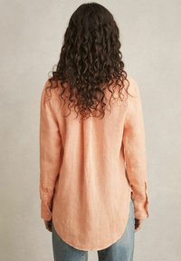 Persikų spalvos lininė marškiniai su išlenktu hemu, ilgomis rankovėmis ir mygtukų detalėmis ant rankogalių, rodomi iš nugaros.