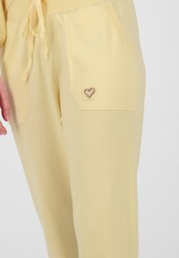 Lichtgele joggers van zacht materiaal, met twee voorkruiszakken en een klein hartvormig metalen accent op de rechterzak.