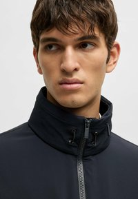 Svart jacka med hög krage, med en framslagningsdragkedja och detalj med snörning. Slät textur med subtil varumärkeslogotyp på dragkedjan.