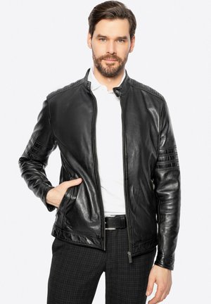 Chaqueta de cuero negra con cierre de cremallera, cuello alzado, detalles acolchados en los hombros y dos bolsillos laterales, llevada sobre una camisa blanca.