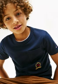 Camiseta de manga corta azul marino con cuello redondo, que presenta un bolsillo en el pecho y un diseño de emblema bordado con un león y acentos de laurel.