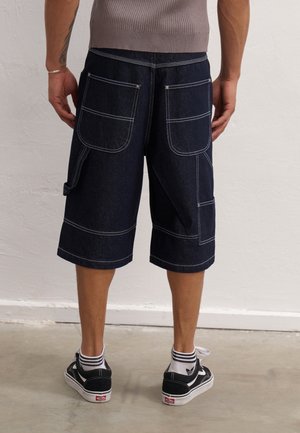 Homme portant un short cargo en denim foncé avec de grandes poches arrière, des chaussettes blanches à rayures et des baskets noires, debout sur un sol en béton.