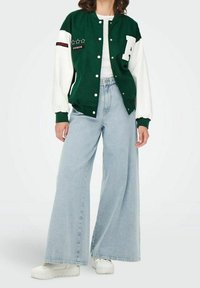 Zielono-biała kurtka sportowa z zapięciem na guziki, zdobiona gwiazdkami i akcentami tekstowymi, połączona z szerokimi jasnobłękitnymi dżinsami i białymi sneakersami.