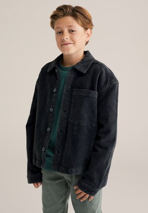 Chaqueta de pana negra con un corte relajado, bolsillo frontal y cierre de botones, combinada con una camisa verde y pantalones verde claro.