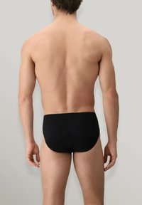 Slip da bagno neri con una texture liscia, un design a vita bassa e copertura minima, mostrati su un modello di spalle contro uno sfondo grigio chiaro.