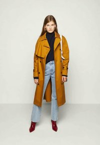 Ženska v gorčično rumenem plašču, črnem polnilu, svetlo modrih kavbojkah, rdečih gležnjarjih in bež torbici, stoji pred enotno ozadjem.