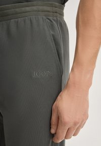 Grau sportliche Hose aus einem strukturierten Stoff, mit einem gerippten Bund und einem gestickten "JOOP!"-Logo auf der Vorderseite. Tasche sichtbar.
