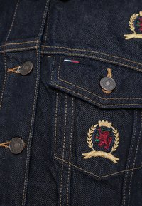 Tmavomodrá rifľová bunda s bronzovým kovaním, náprsné vrecká a vyšívaný znak s levom, zdobený zlatými a zelenými detailmi.