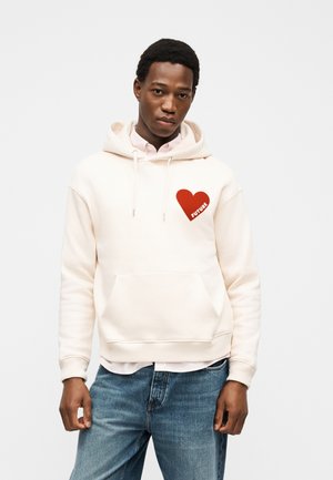 Jeune homme portant un sweat à capuche crème avec un cœur rouge et le texte « FUTURE », associé à un jean bleu clair, debout devant un fond blanc uni.