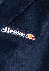 Tamsiai mėlyna poliesterio medžiaga su minkšta tekstūra, turinti baltą "ellesse" logotipą ir spalvotą pusiau saulės emblemą oranžinės ir raudonos spalvų.