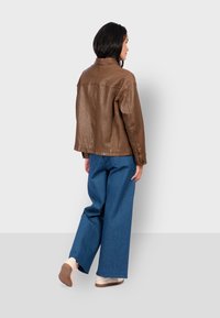 Giacca di pelle marrone con una vestibilità comoda, caratterizzata da un collo a camicia. Abbinata a ampi pantaloni in denim blu e scarpe chiare.
