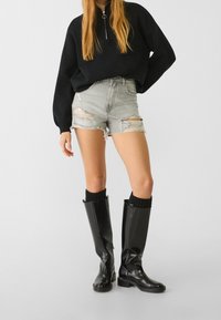Persona vestida con un suéter negro de cremallera, pantalones cortos de denim claro rasgados y botas altas negras con calcetines negros.