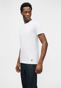 Άνδρας που φοράει λευκό κοντομάνικο μπλουζάκι με τσέπη στο στήθος και μικρή κεντητή λεπτομέρεια κοντά στο τελείωμα, συνδυασμένο με σκούρο μπλε τζιν.