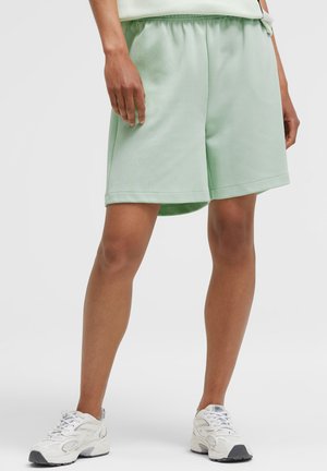 Person trägt hellgrüne knielange Shorts und weiße Laufschuhe, steht vor einem einfarbigen hellen Hintergrund.