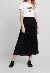 Černá plisovaná midi sukně s volným designem, zkombinovaná s bílým grafickým tričkem s černým textem a ikonou oranžové včelí plástve, nošena s černými teniskami.