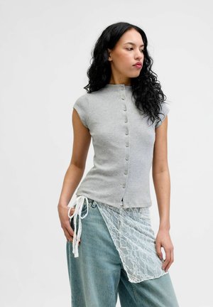 Femeie cu păr lung și creț, purtând un top gri fără mâneci cu nasturi și blugi albaștri, și o șorț alb din dantelă legat în talie.