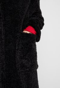 Svart, fluffig morgonrock med mjuk textur. Har sidfickor och en synlig röd ärm under. Designen är lång och lös.