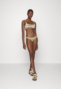 Biquíni listrado em verde, branco e preto, com um top estilo bralette e calções de corte baixo. Modelo combina com sandálias de cores claras.