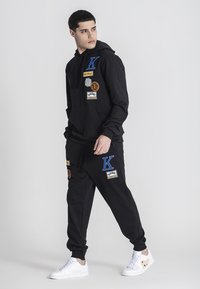 Felpa nera con cappuccio e pantaloni della tuta in misto cotone, caratterizzati da toppe colorate e ricami. Indossati con sneakers bianche. Sfondo semplice.