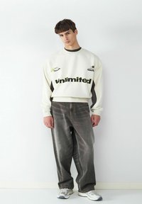 Jasno beżowy sweter z napisem "Unlimited", ciemne akcenty, w połączeniu z luźnymi szarymi dżinsami i szarymi trampkami. Casuowy design streetwear.