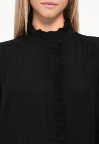Svart bluse med høy krage og volang i blonder, frontåpning med knapper og glatt, strukturert stoff.