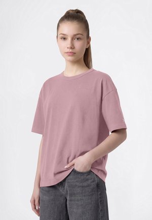 Ung kvinde med håret samlet tilbage iført en løs pink T-shirt og grå jeans, stående med den ene hånd i lommen mod en ensfarvet baggrund.