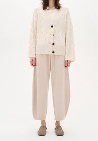 Beige kabelstickad cardigan med stora svarta knappar, i kombination med lösa, ljusbeige byxor och gråa slip-on skor. Mjuka texturer genom hela outfiten.