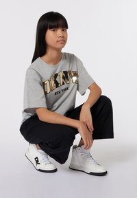 T-shirt gris avec le logo doré "DKNY", pantalons noirs et baskets blanches. Le mannequin est accroupi avec une expression neutre devant un fond uni.