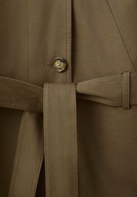 Gros plan d'un manteau en tissu kaki avec un bouton et une ceinture en tissu attachée à travers une boucle à la taille.