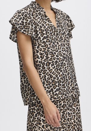 Kvinde, der har en bluse med korte ærmer og et nederdel med et brunt, sort og hvidt leopardmønster mod en ensfarvet baggrund.