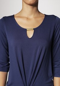 Marinblå topp med rundad halsringning, prydd med ett guldackcent vid halsringningen och rynkad detalj vid midjan, tillverkad av mjukt tyg.