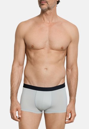 Uomo con barba curata che indossa boxer grigio chiaro con fascia in vita nera, in piedi davanti a uno sfondo bianco uniforme.