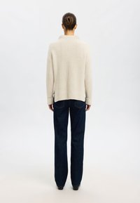Cremfarbener gerippter Strickpullover mit hohem Kragen, kombiniert mit weit geschnittenen, dunklen Jeans. Schlichtes Design, ohne sichtbare Muster oder Verzierungen.