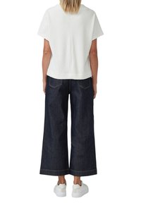 Weißes strukturiertes Kurzarmoberteil kombiniert mit lockeren, dunklen Denim-Hosen im weit geschnittenen Stil. Das Outfit wird mit weißen Sneakern mit dezenten Akzenten abgerundet.