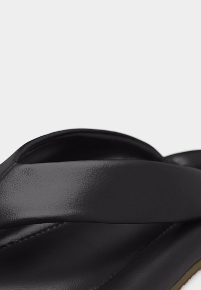 Detailní záběr černého koženého pásku sandálu, který přechází přes polstrovanou stélku s viditelným šitím a texturovaným okrajem podrážky.