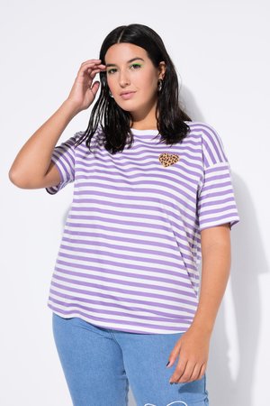 Kobieta o ciemnych, sięgających do ramion włosach, ubrana w fioletowo-białą pasiastą koszulę z łatą w kształcie serca w panterkę oraz niebieskie dżinsy.