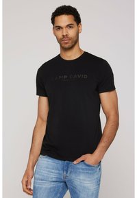 Camiseta de algodón negra con mangas cortas, cuello redondo y el logo "CAMP DAVID" en verde oscuro en la parte delantera, combinado con vaqueros azules.