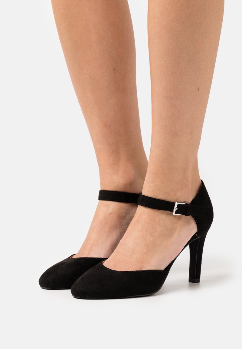 Chaussures à talons hauts en daim noir avec brides à la cheville et boucles argentées portées pieds nus sur un fond clair uni.